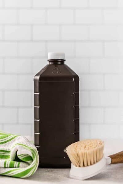 Bottle of hydrogen peroxide in front of a white subway tile wall with green stripe kitchen towel and cleanign brush