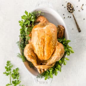 whole chicken with brown crispy skin surrounded by fresh herbs on a white background