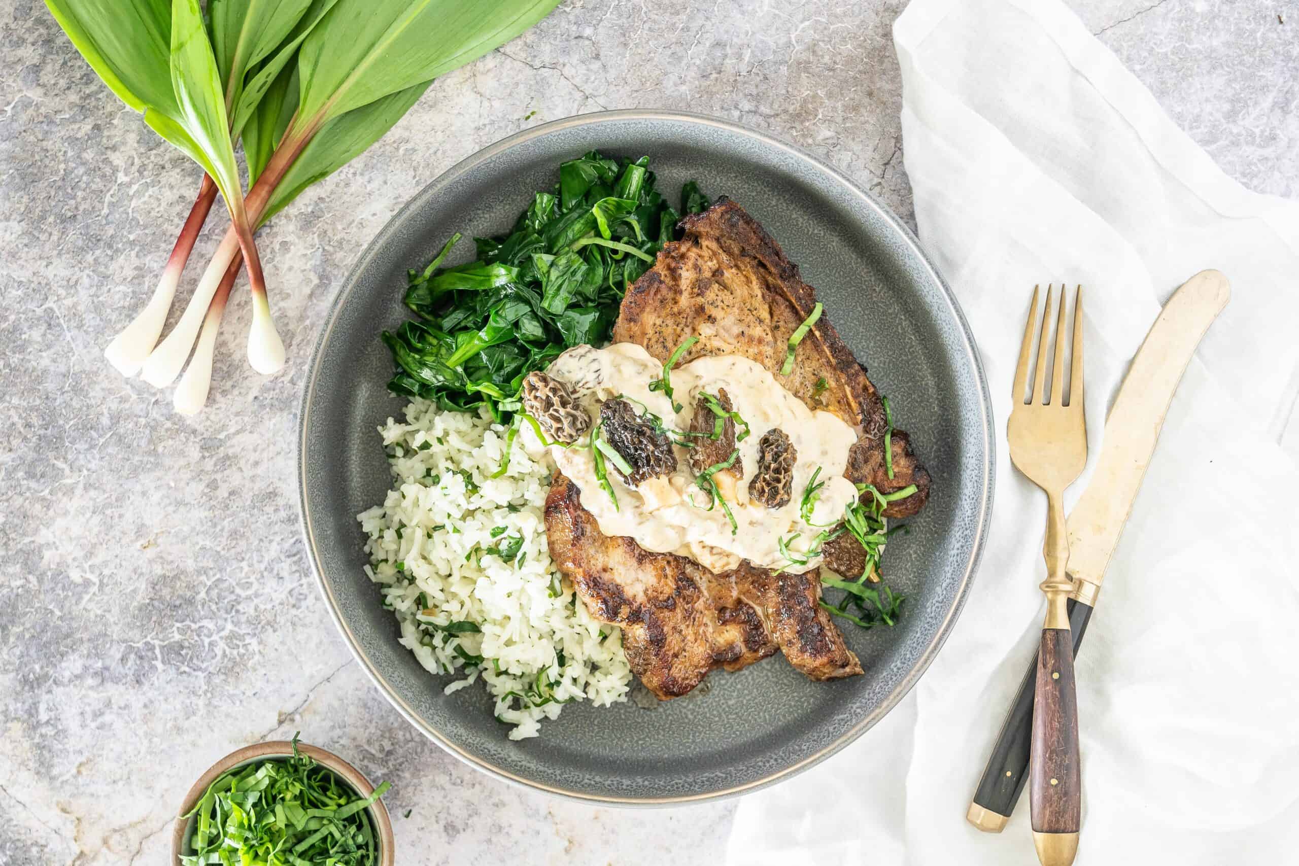 gray background with a gray bowl, holding sautéed green, rice and pork chops topped with a parmesan sauce with ramps and morels