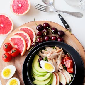 A vibrant breakfast flat lay featuring a mix of fruits, avocado, eggs, tomatoes, and sliced meats on a wooden platter.