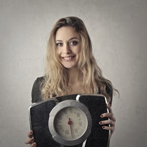 Young woman with blond hair smiling, holding a vintage bathroom scale indoors.