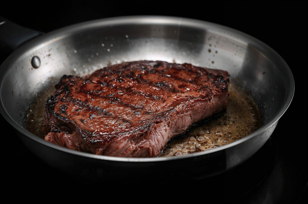 Close up view of a stainless steel skillet with a seared crust on a thick ribeye steak cooking on a burner against a dark backdrop