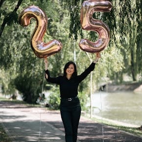 Happy woman in black outfit celebrating her 25th birthday with balloons in a sunny park.