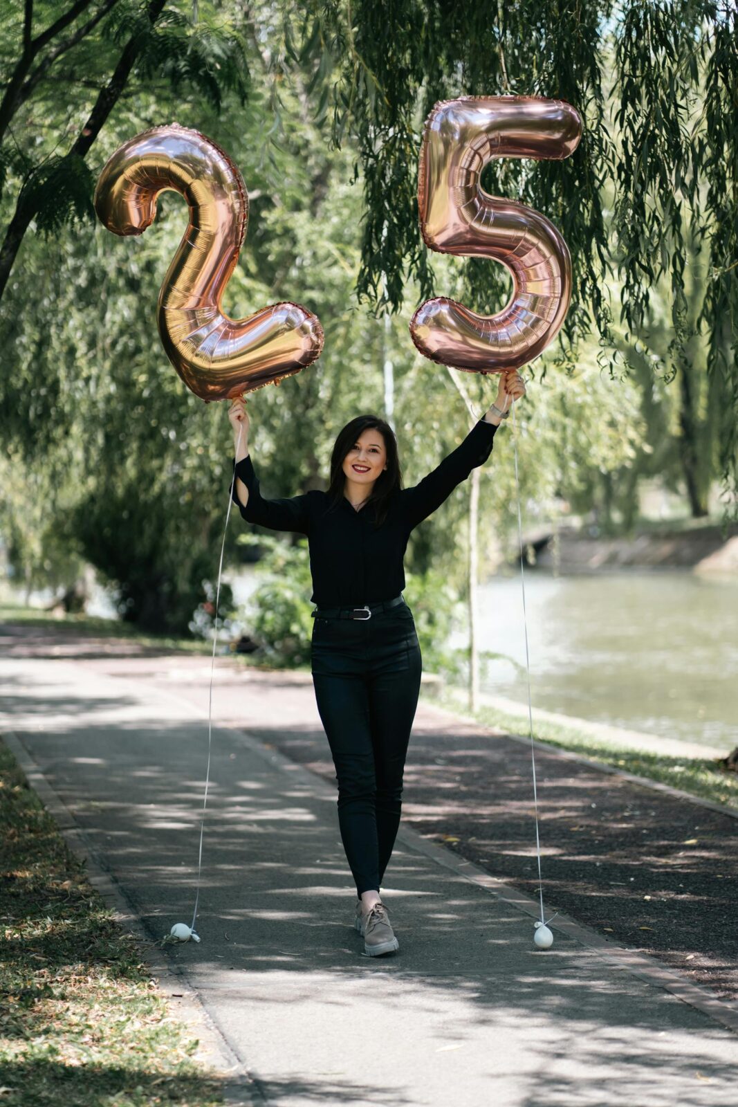 Happy woman in black outfit celebrating her 25th birthday with balloons in a sunny park.