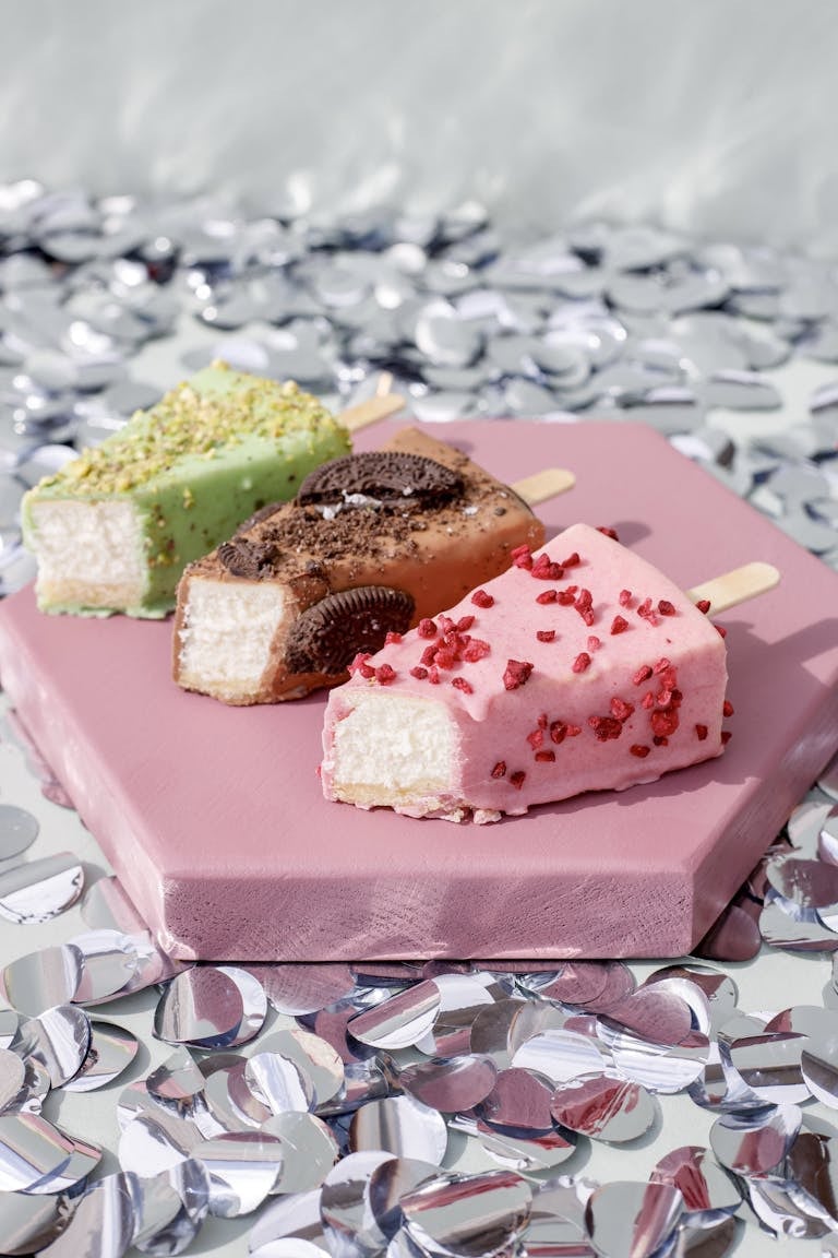 Vibrant assorted ice cream bars with toppings on a pink tray, surrounded by confetti.