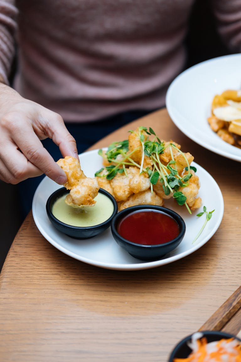 Close-up of a hand dipping crispy tofu bites into sauces, garnished with microgreens.