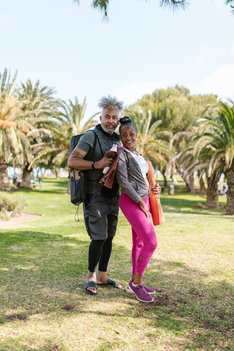 Senior couple posing with yoga mats in a sunny park, ready for fitness activity.