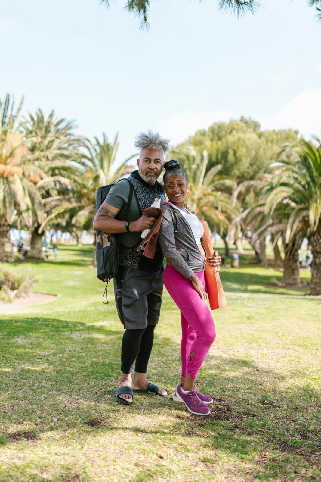 Senior couple posing with yoga mats in a sunny park, ready for fitness activity.