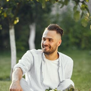 Smiling man sitting on grass with a basket of fresh vegetables in a sunny garden.