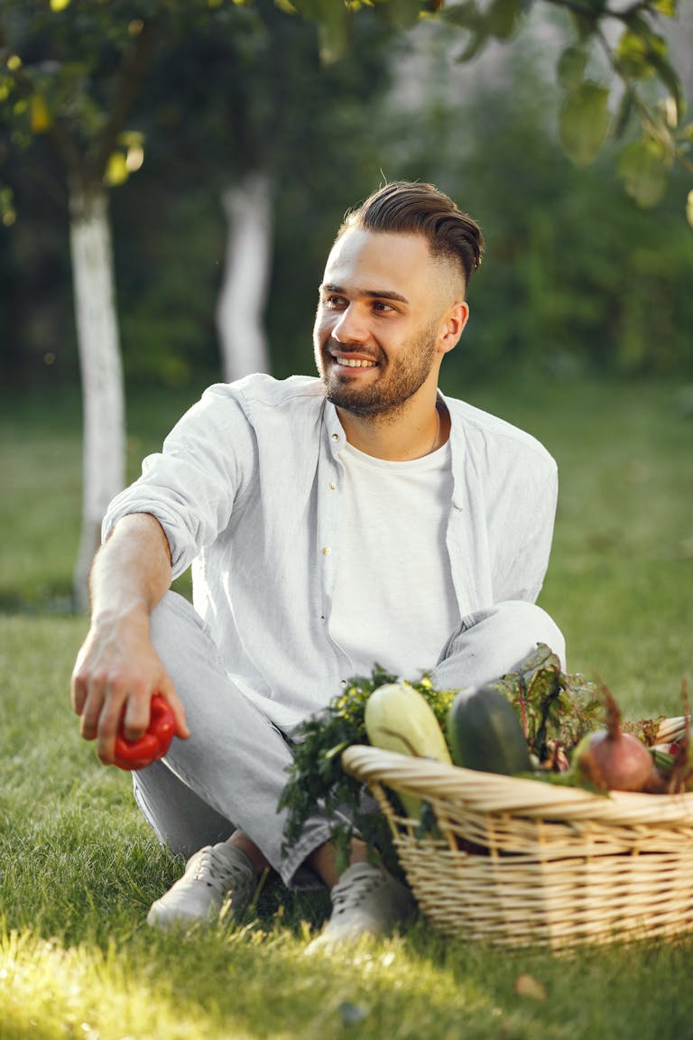 Smiling man sitting on grass with a basket of fresh vegetables in a sunny garden.