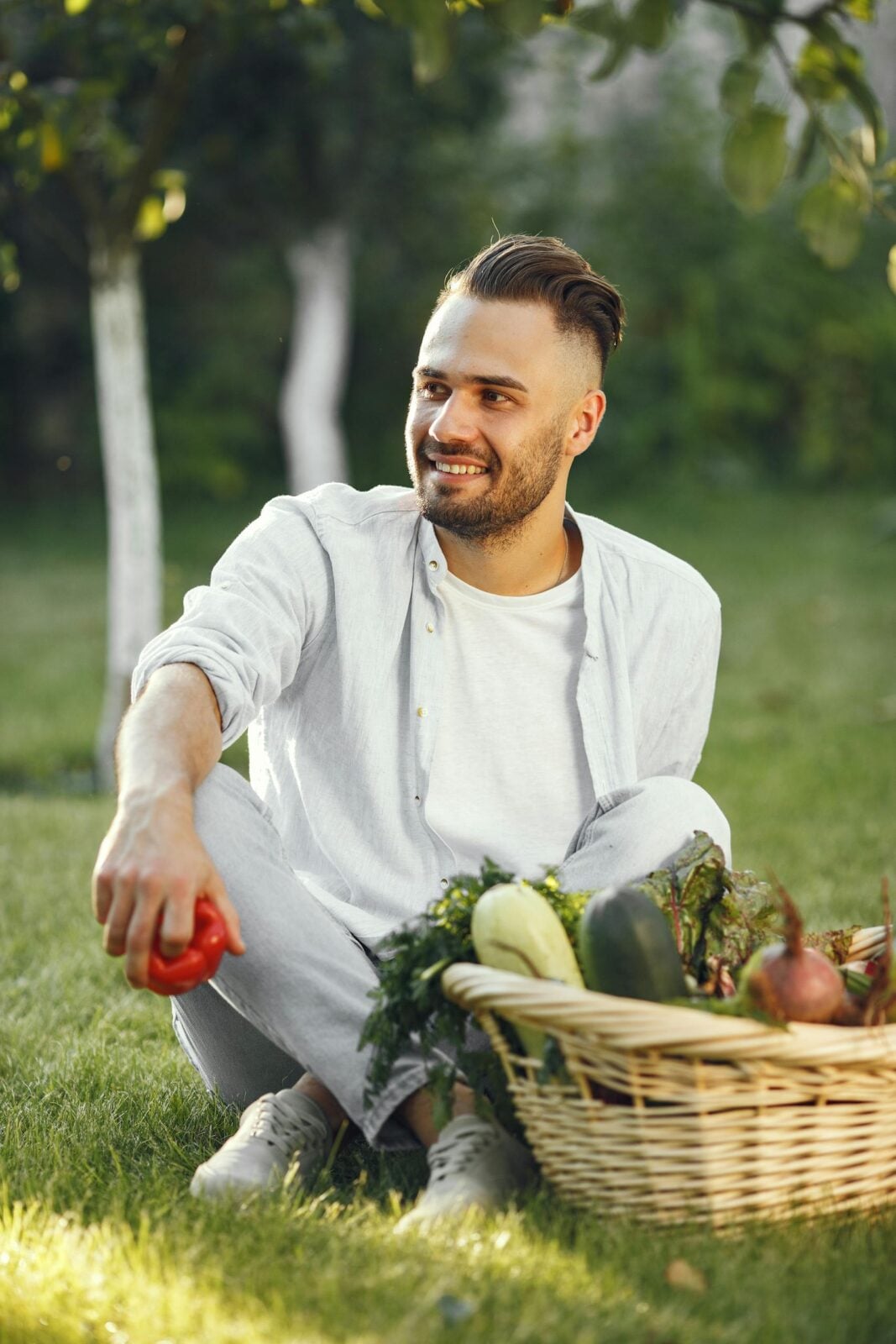Smiling man sitting on grass with a basket of fresh vegetables in a sunny garden.