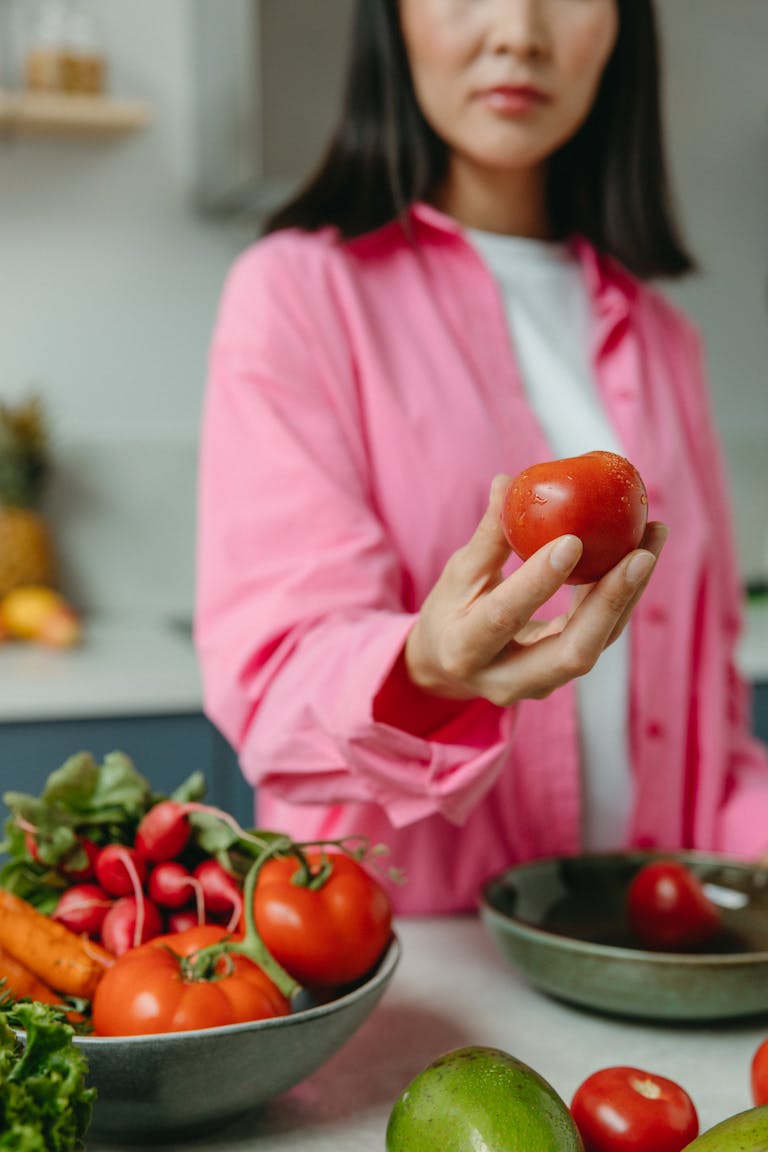 A person holding a fresh tomato over a bowl of assorted vegetables in a kitchen setting.