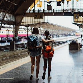 Two young backpackers embracing at a bustling train station, ready for an adventure.