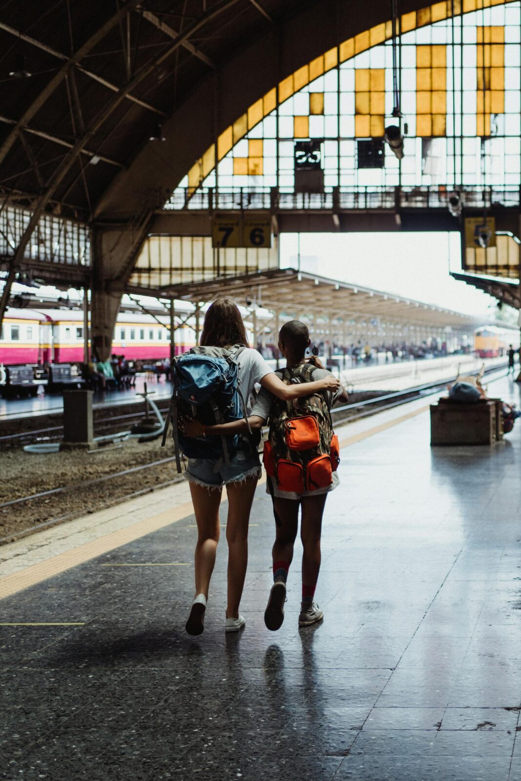 Two young backpackers embracing at a bustling train station, ready for an adventure.