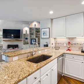 A modern kitchen with granite countertops opens into a stylish living room, featuring elegant furniture and built-in shelves.