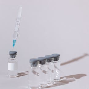 A syringe with a clear substance is placed into a glass vaccine vial on a white surface.