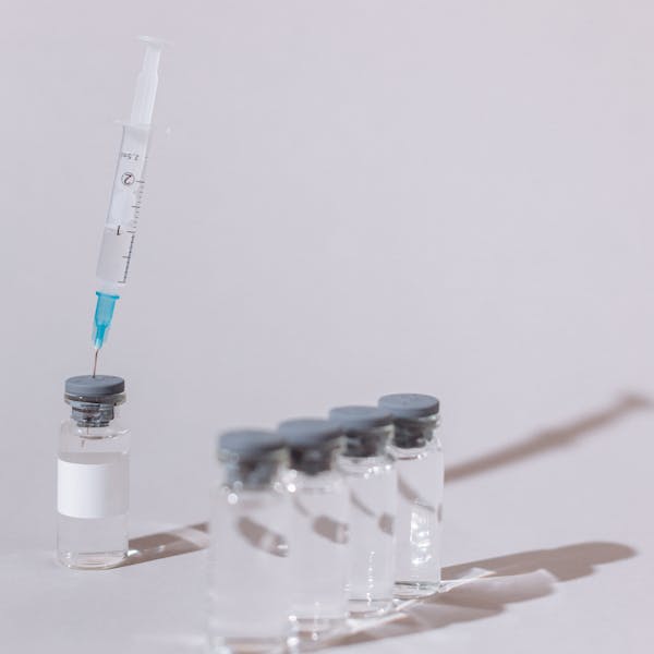 A syringe with a clear substance is placed into a glass vaccine vial on a white surface.