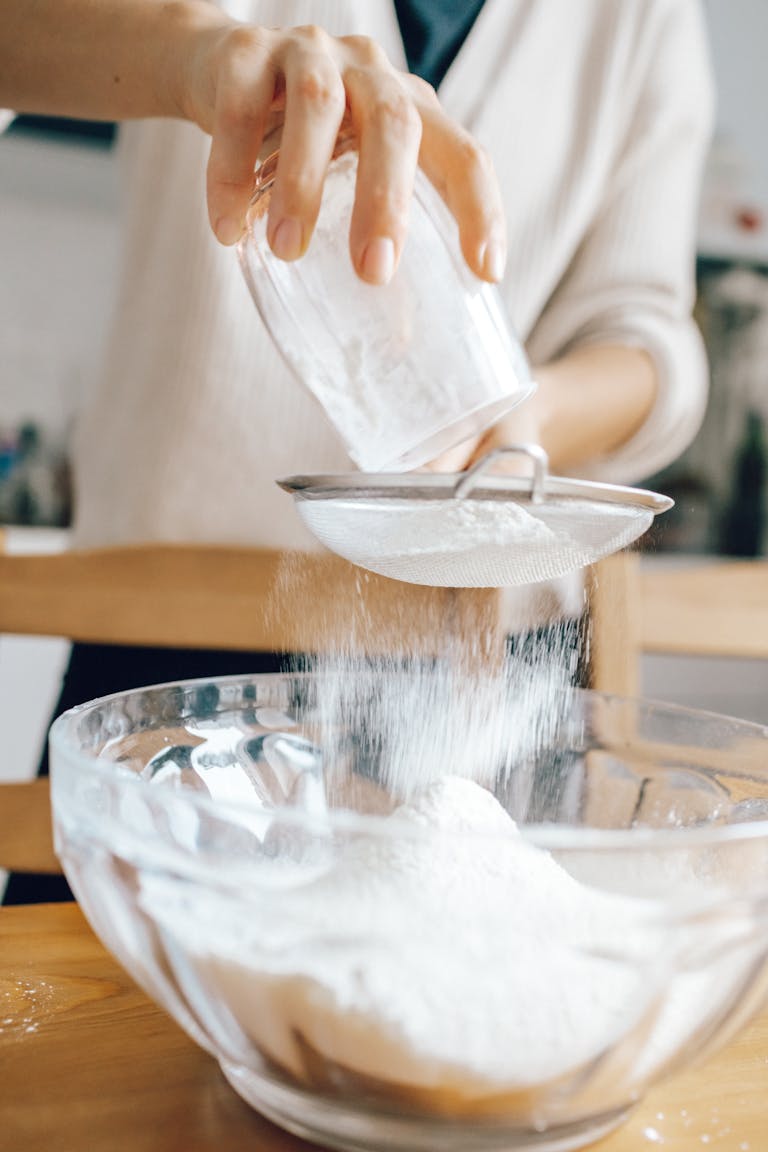 Close-up of hands sifting flour into a glass bowl, ideal for baking tutorials or food blogs.