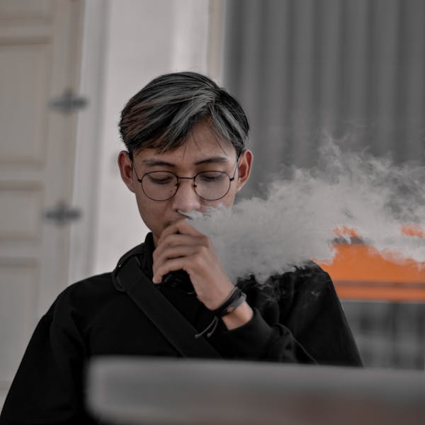 Portrait of a young adult wearing glasses while vaping indoors, producing a visible cloud of vapor.
