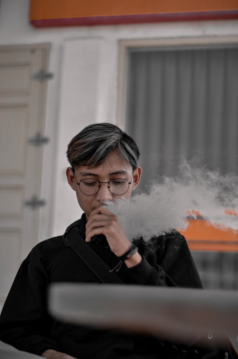 Portrait of a young adult wearing glasses while vaping indoors, producing a visible cloud of vapor.