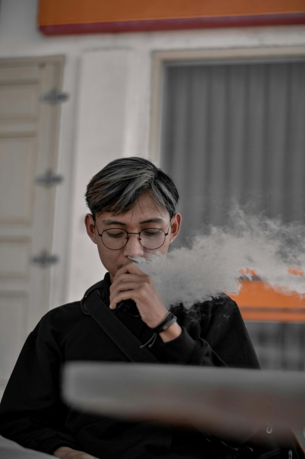 Portrait of a young adult wearing glasses while vaping indoors, producing a visible cloud of vapor.