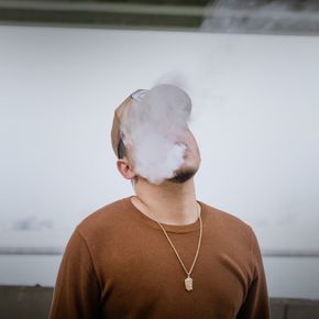 Man in brown sweater and cap exhaling vape smoke outdoors.