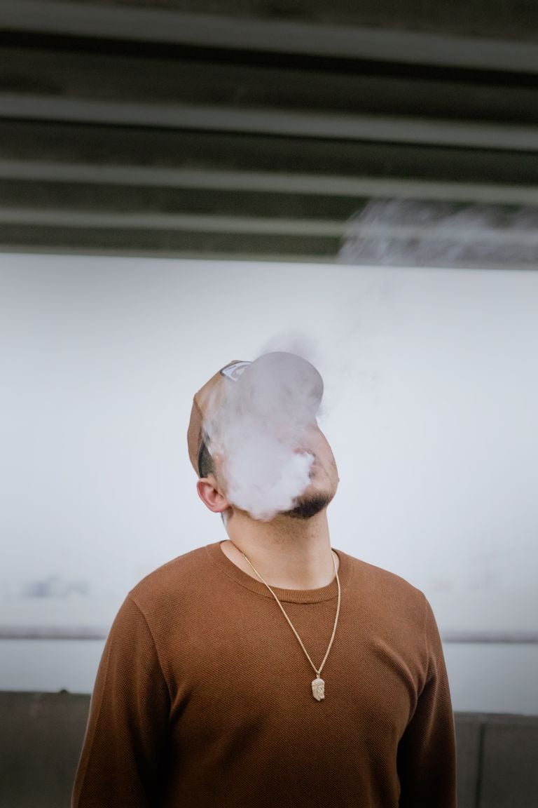 Man in brown sweater and cap exhaling vape smoke outdoors.