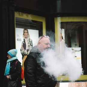 A man in a jacket vapes on a bustling city sidewalk, creating a cloud of smoke.