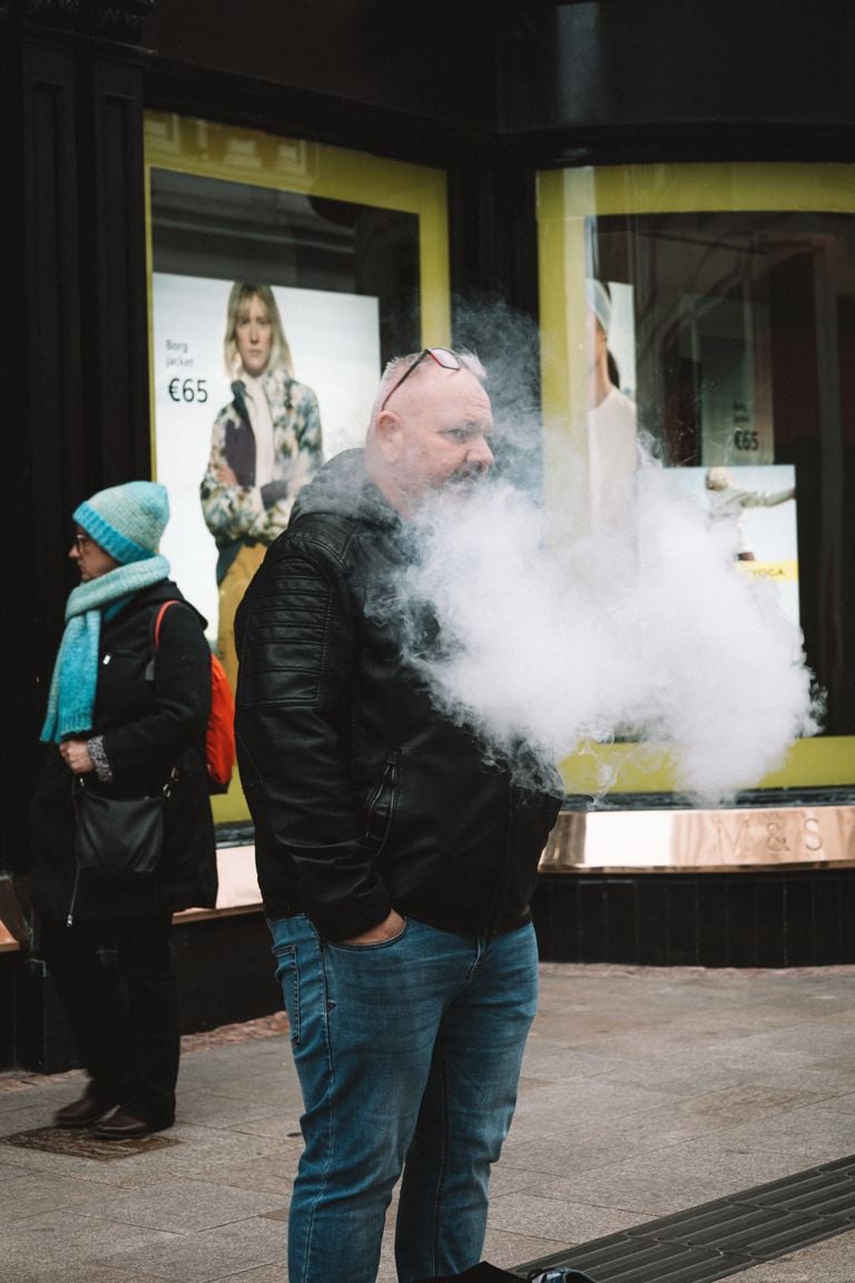 A man in a jacket vapes on a bustling city sidewalk, creating a cloud of smoke.