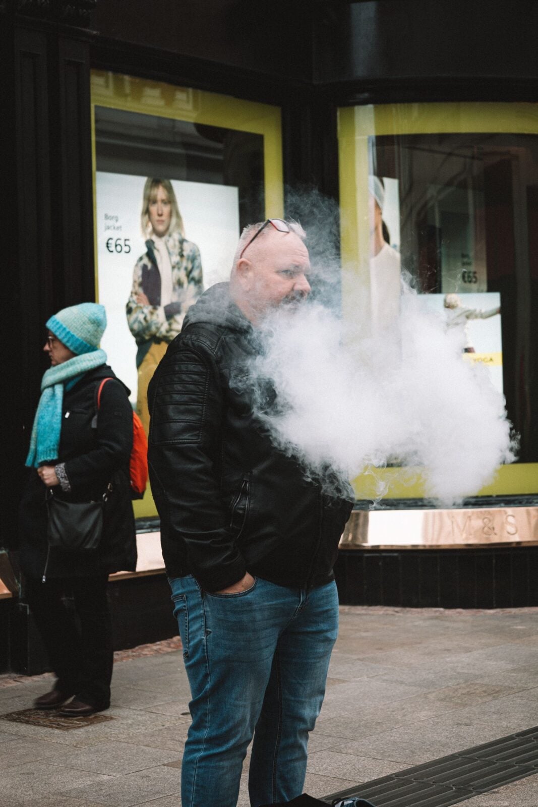 A man in a jacket vapes on a bustling city sidewalk, creating a cloud of smoke.