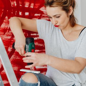 A woman on a ladder uses a drill on an art project with a red abstract background.