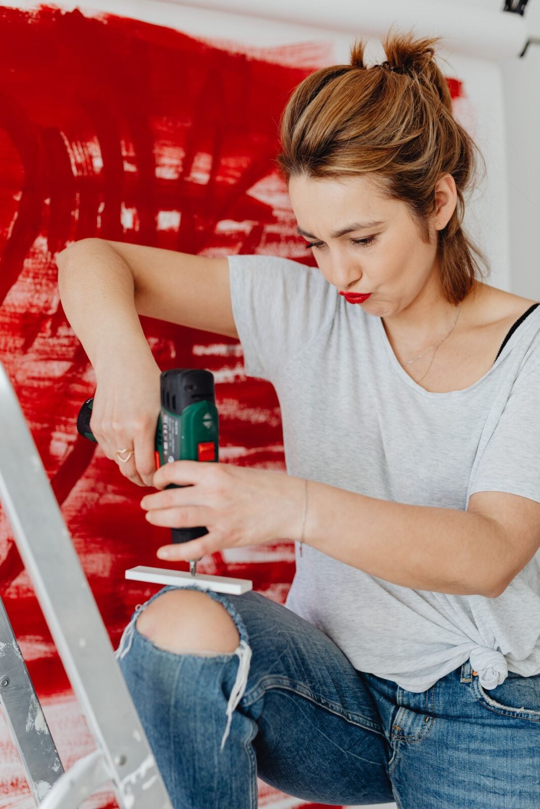 A woman on a ladder uses a drill on an art project with a red abstract background.