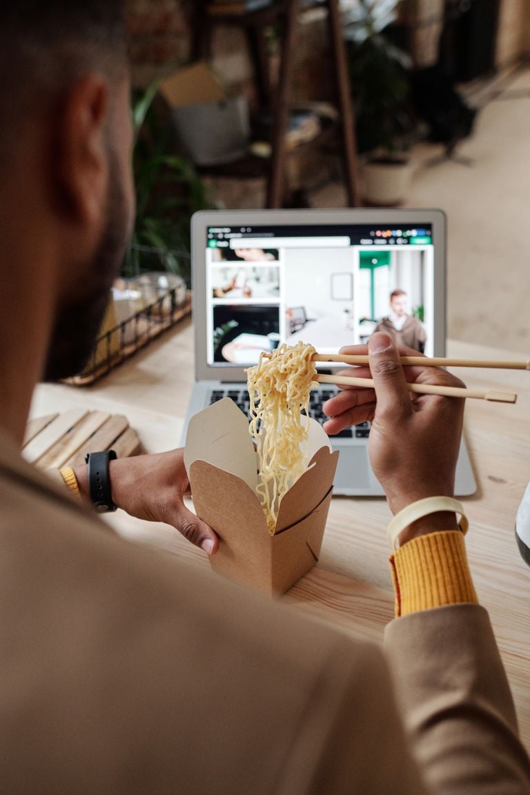A person eating noodles with chopsticks while using a laptop, indoors.