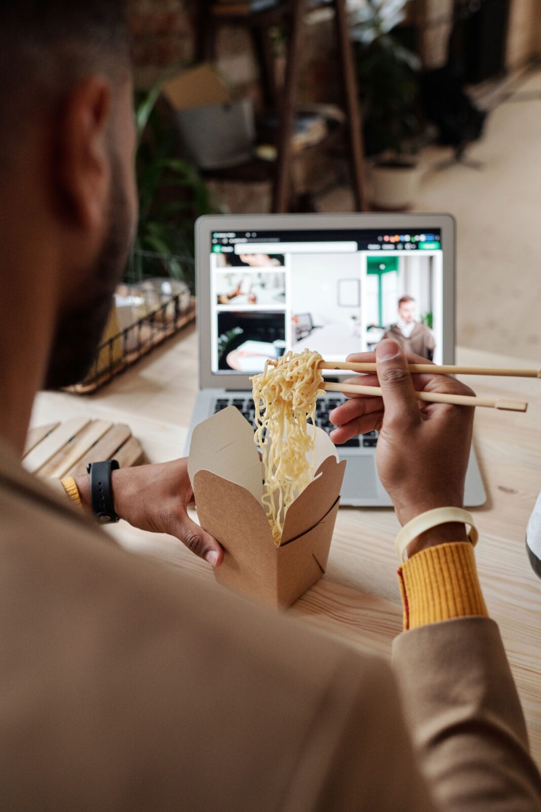 A person eating noodles with chopsticks while using a laptop, indoors.