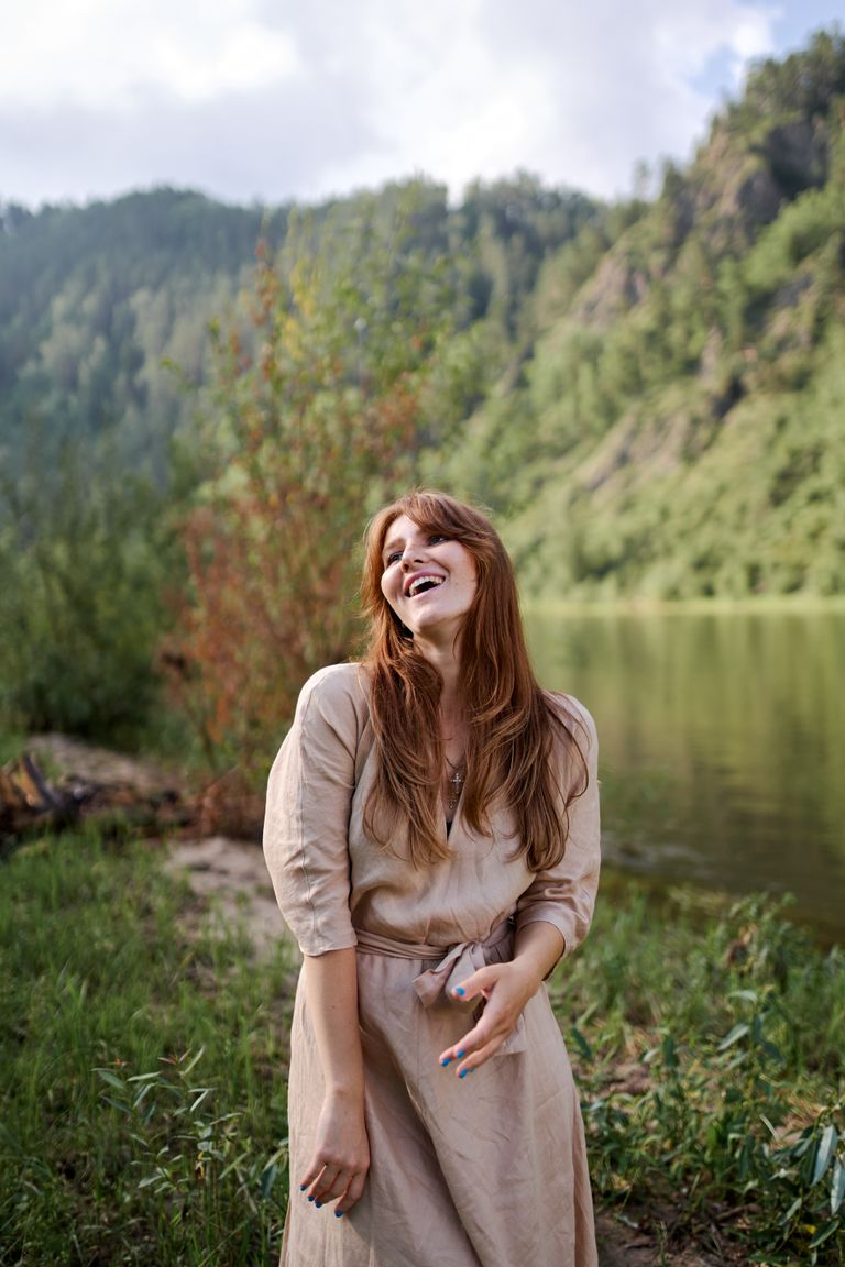Young cheerful female in casual dress looking up near river and mount with lush green trees in summer