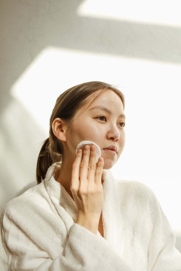 Woman in a robe applying skincare products indoors with natural lighting.