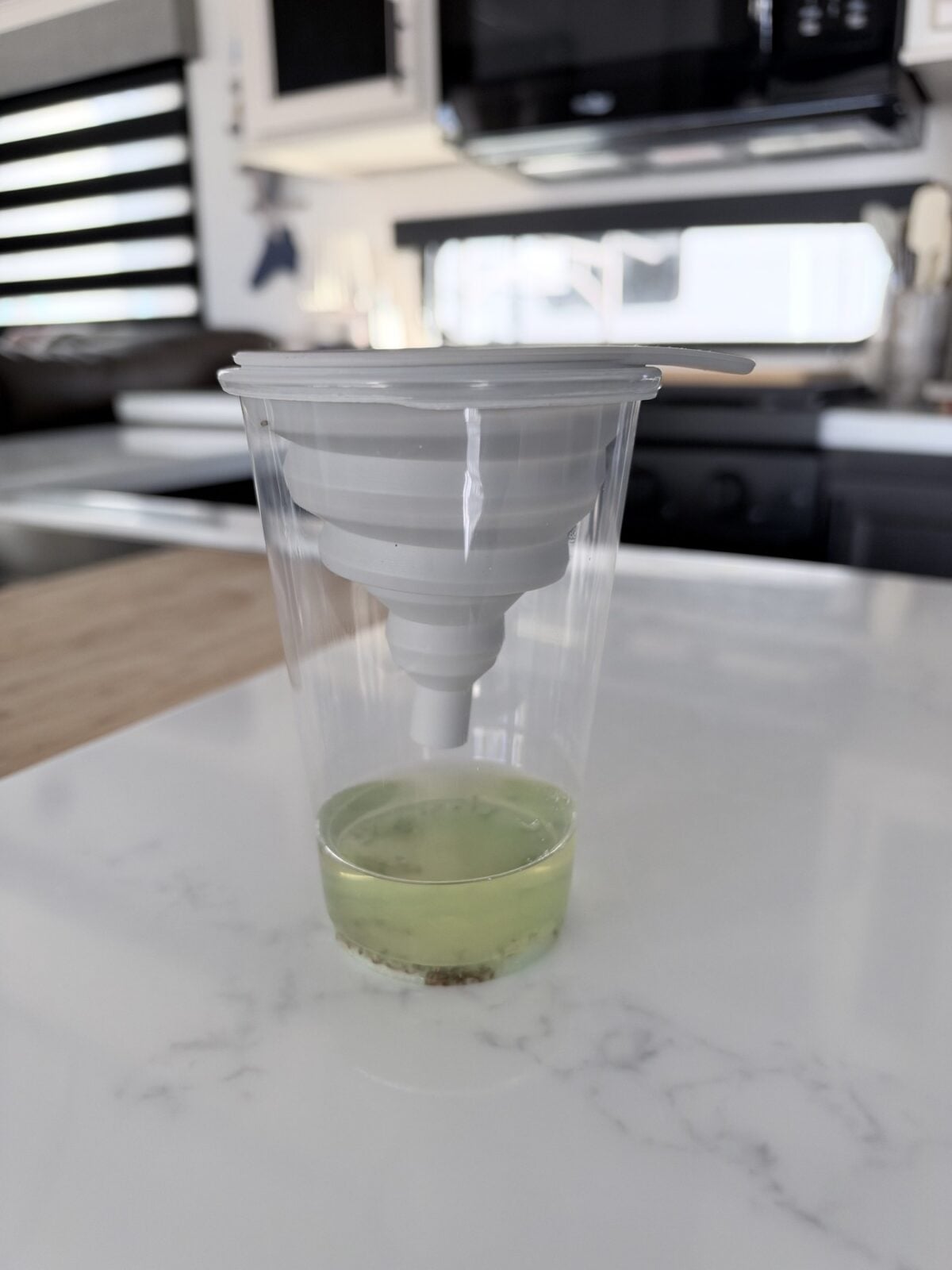 Glass holding apple cider vinegar and dish soap beneath a white collapsible silicone funnel